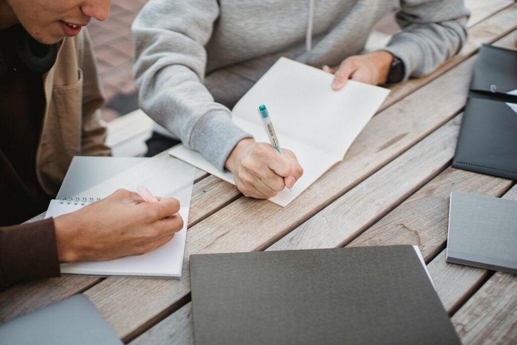 Die Kunst der Hausarbeit Fragestellung: Tipps und Tricks für deine wissenschaftliche Arbeit 5 High angle crop male students in casual clothes taking notes in copybooks while working on home assignment together in park